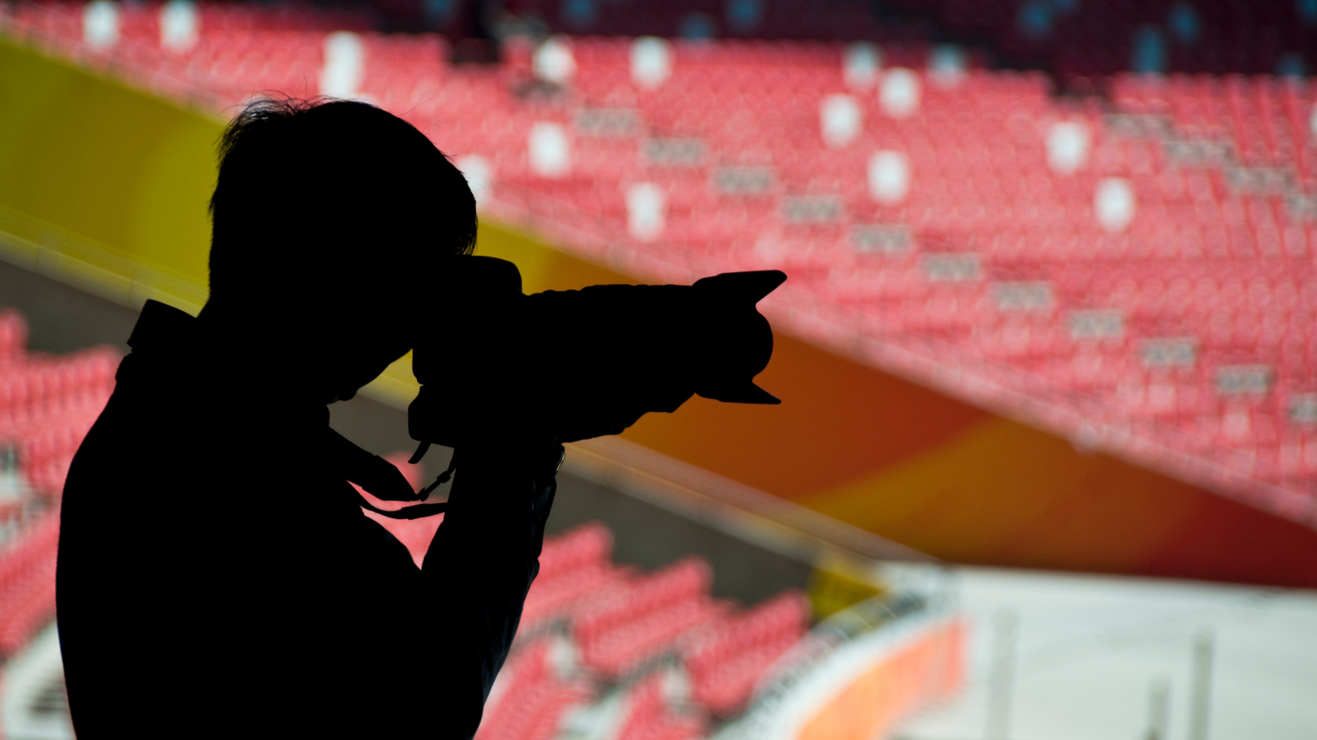 A silhouette of a man with a professional filming camera points towards the centre of a sporting arena as empty audience seating appear behind him.